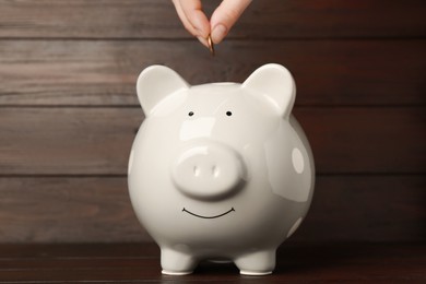 Photo of Woman putting coin into piggy bank at wooden table, closeup