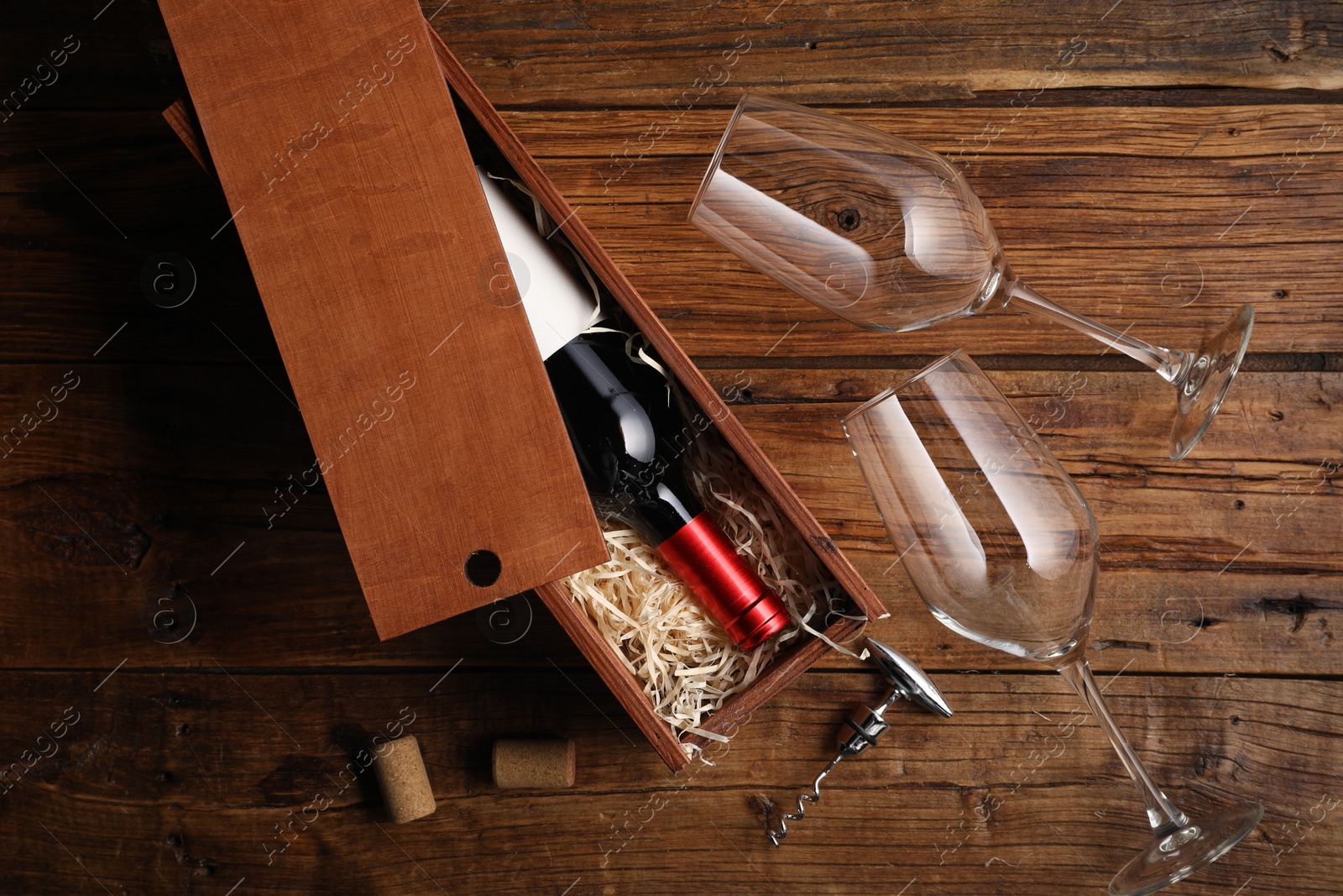 Photo of Box with wine bottle, glasses, corkscrew and corks on wooden table, flat lay