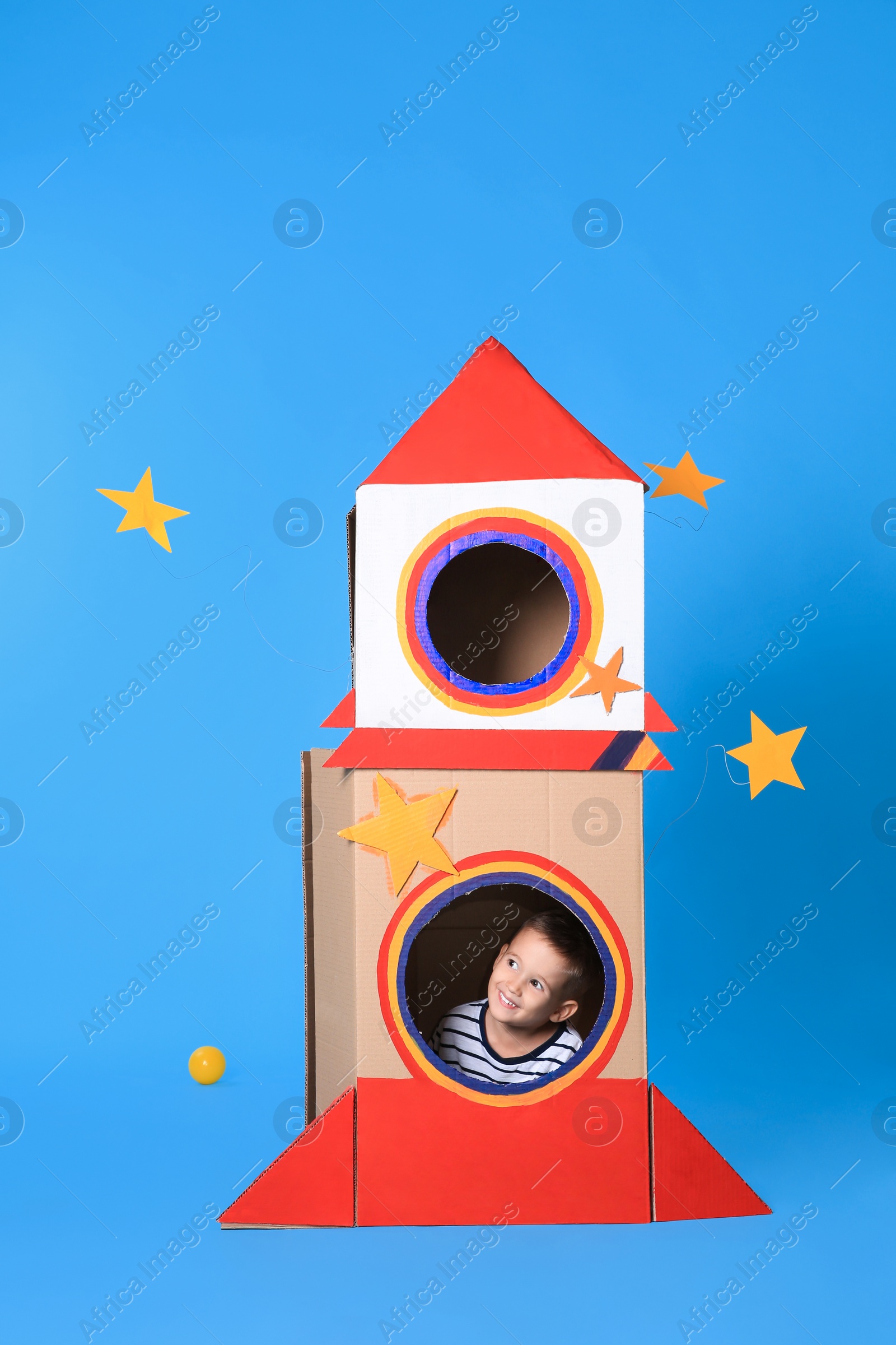 Photo of Cute little boy playing with cardboard rocket on light blue background