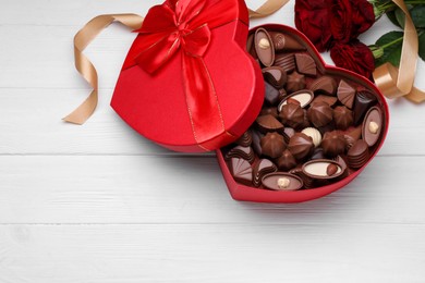 Photo of Heart shaped box with delicious chocolate candies, roses and ribbon on white wooden table, flat lay. Space for text