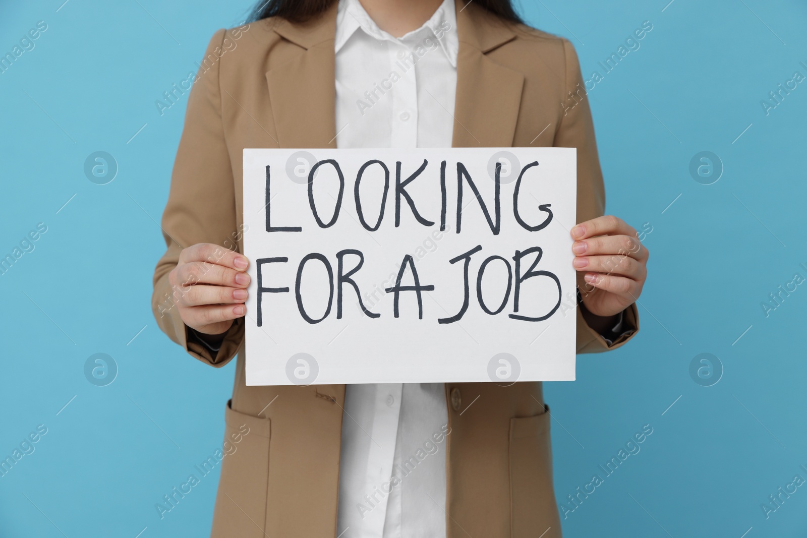 Photo of Young unemployed woman holding sign with phrase Looking For A Job on light blue background, closeup