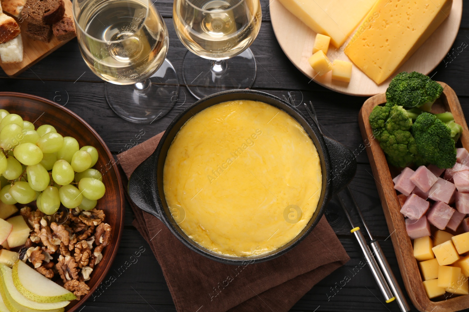 Photo of Fondue pot with melted cheese, glasses of wine and different products on black wooden table, flat lay