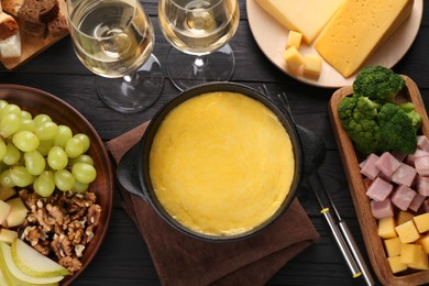 Fondue pot with melted cheese, glasses of wine and different products on black wooden table, flat lay