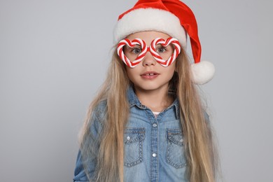 Girl wearing decorative Christmas eyeglasses in shape of hearts and Santa hat on light grey background