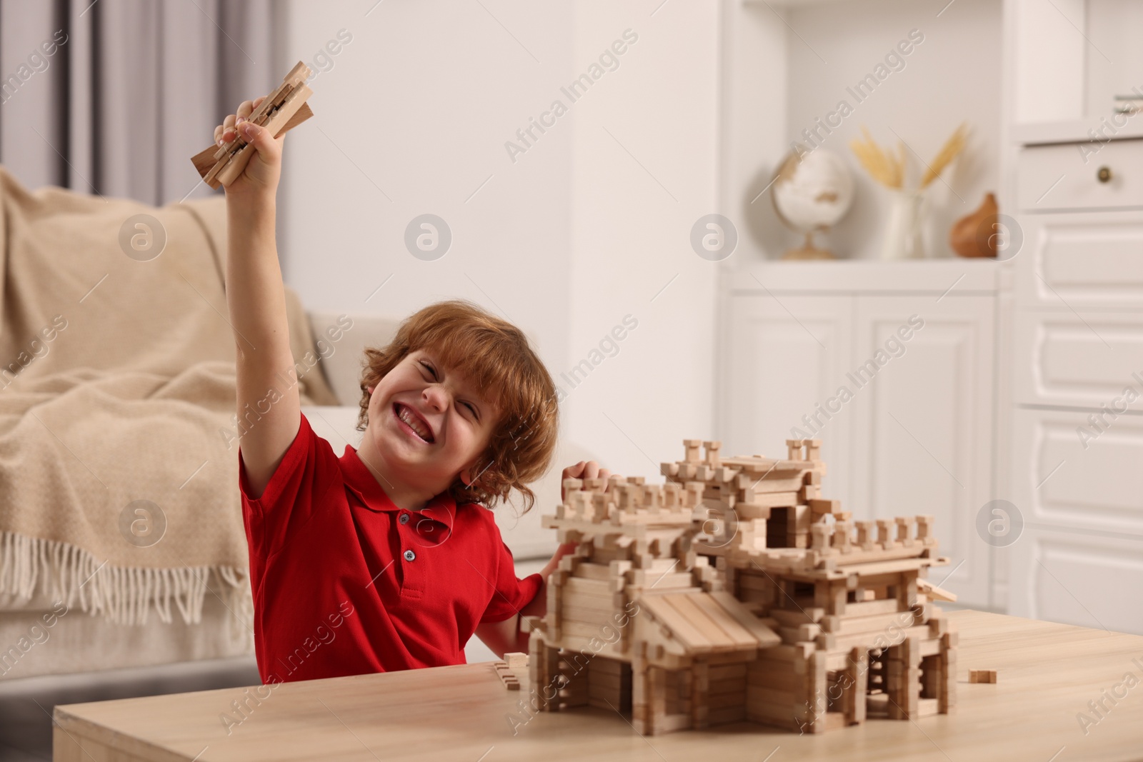 Photo of Emotional boy playing with wooden castle at table in room. Child's toy