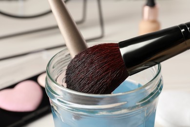 Photo of Cleaning makeup brushes in jar with special liquid on white table, closeup