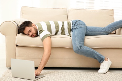 Photo of Lazy young man using laptop while lying on sofa at home