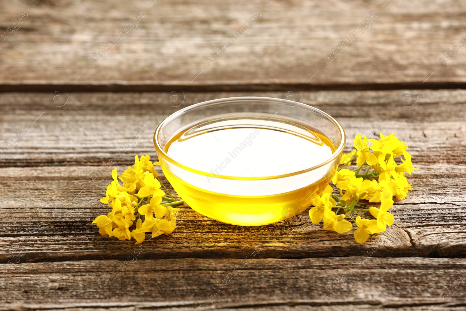 Photo of Rapeseed oil in glass bowl and beautiful yellow flowers on wooden table