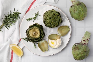 Photo of Delicious cooked artichokes with tasty sauce served on white tiled table, flat lay