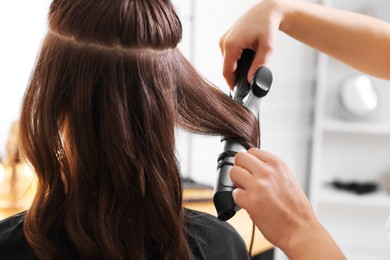 Hairdresser using curling hair iron while working with woman in salon, closeup