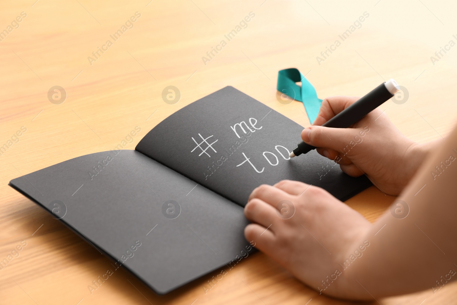 Photo of Woman writing hashtag MeToo in notebook near teal awareness ribbon at table, closeup. Stop sexual assault