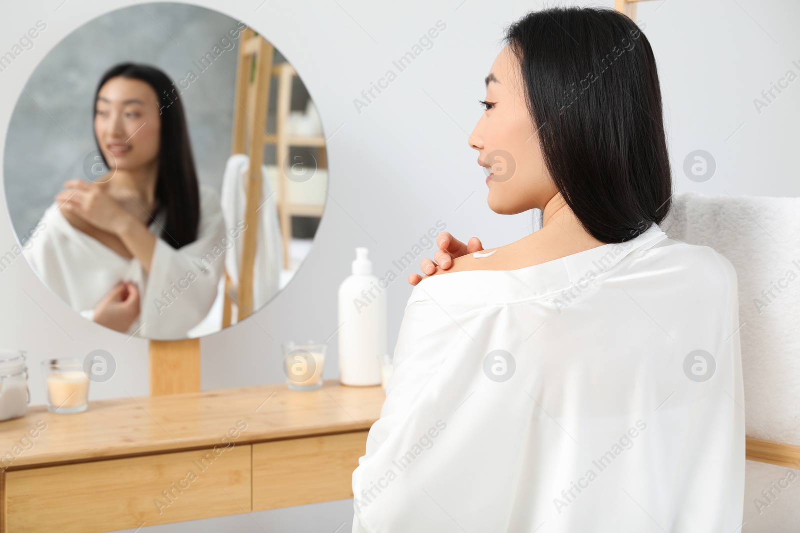 Photo of Beautiful young Asian woman applying body cream on shoulder in bathroom