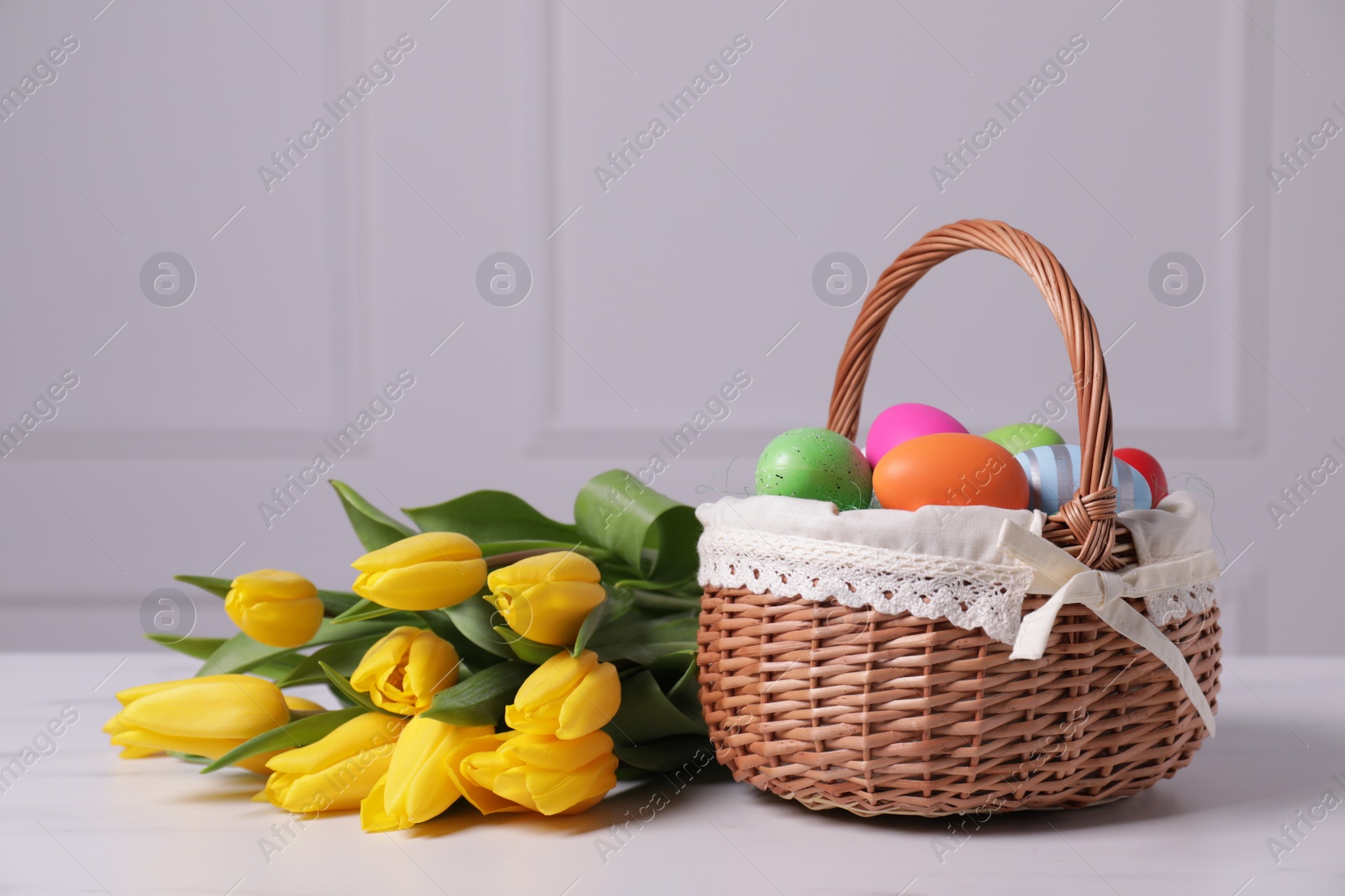 Photo of Easter basket with painted eggs near bouquet of tulips on white table. Space for text
