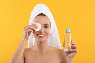 Smiling woman removing makeup with cotton pad and holding bottle on yellow background