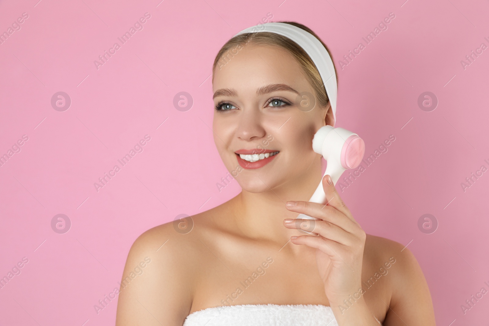 Photo of Young woman washing face with cleansing brush on pink background. Cosmetic product