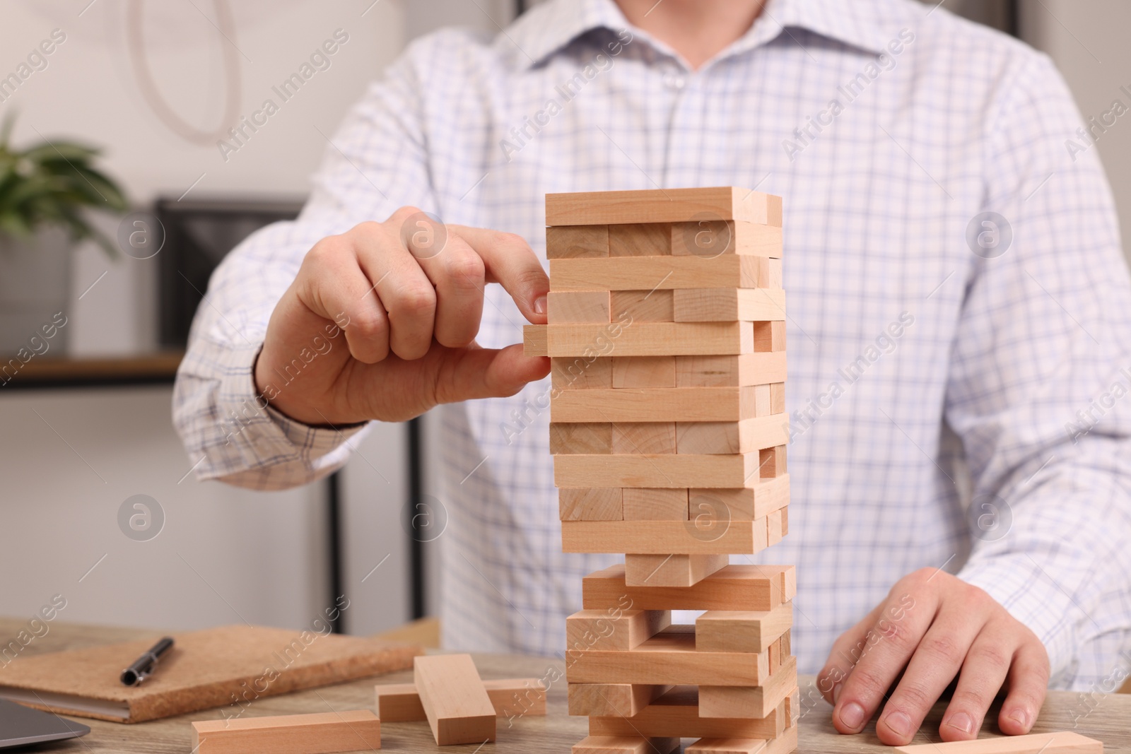 Photo of Playing Jenga. Man removing wooden block from tower at table indoors, closeup