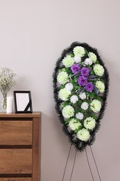 Wreath of plastic flowers and frame with black ribbon, burning candle on commode in room. Funeral attributes