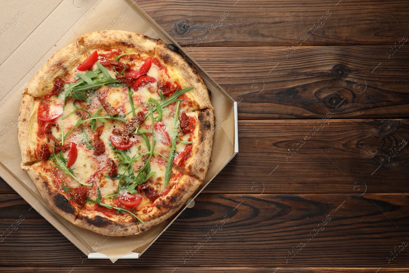 Photo of Tasty pizza with cheese, tomato and arugula in cardboard box on wooden table, top view. Space for text