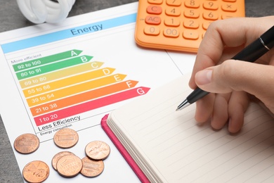 Photo of Woman with pen, notebook, calculator, coins and energy efficiency rating chart at table, closeup