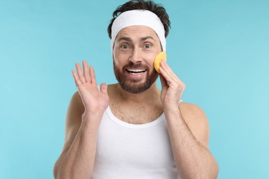 Photo of Man with headband washing his face using sponge on light blue background
