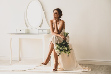 Young bride in wedding dress with beautiful bouquet near mirror indoors