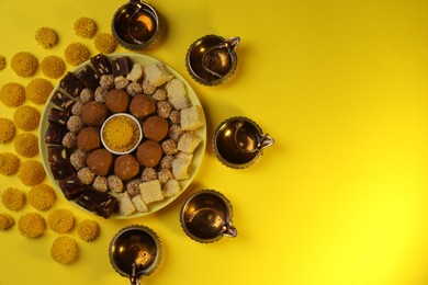 Happy Diwali. Flat lay composition with diya lamps, chrysanthemum flowers and delicious Indian sweets on yellow table. Space for text