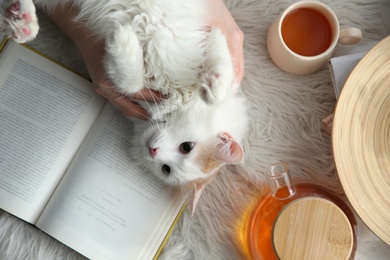 Woman with cute fluffy cat, tea and book on faux fur, top view