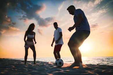 Image of Friends playing football on beach during sunset, low angle view