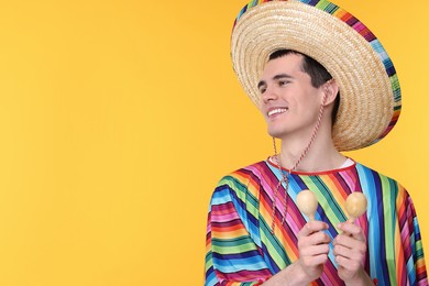 Photo of Young man in Mexican sombrero hat and poncho with maracas on yellow background. Space for text