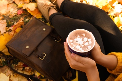 Photo of Woman holding cup of hot drink in park with fallen leaves, closeup view. Autumn season