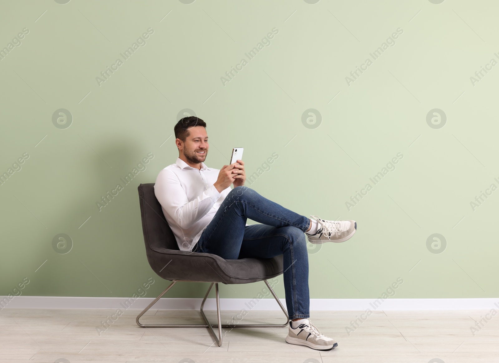 Photo of Happy man sitting in armchair and using smartphone near light green wall indoors