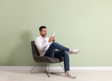 Photo of Happy man sitting in armchair and using smartphone near light green wall indoors