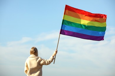 Photo of Woman holding bright LGBT flag against blue sky, back view