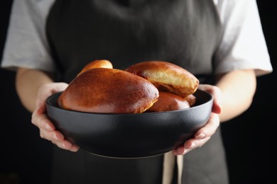 Photo of Woman holding bowl with delicious baked pirozhki on black background, closeup