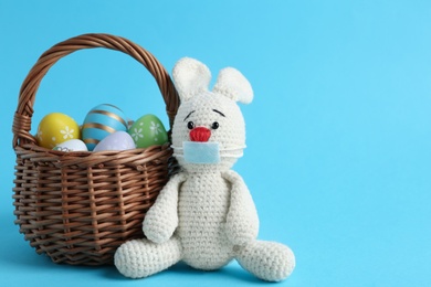 Photo of Toy bunny in protective mask near wicker basket with eggs on light blue background, space for text. Easter holiday during COVID-19 quarantine