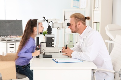 Photo of Children's doctor examining little girl with ophthalmic equipment in clinic