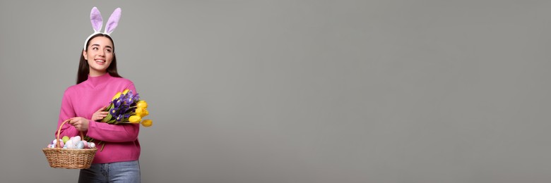 Image of Happy woman with bunny ears holding bouquet and basket full of Easter eggs on light grey background, space for text. Banner design