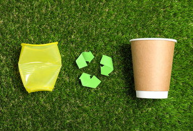 Photo of Recycling symbol, plastic and paper cups on green grass, flat lay