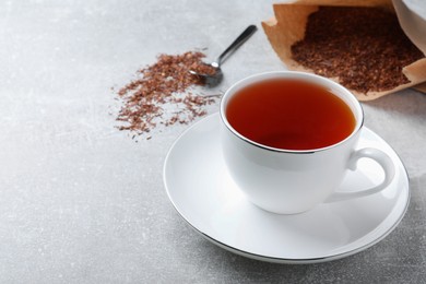 Photo of Ceramic cup of aromatic rooibos tea, spoon and scattered dry leaves on light grey table. Space for text