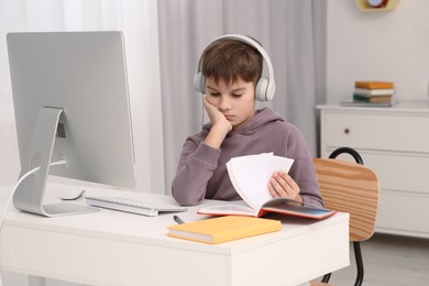 Boy reading book near computer at desk in room. Home workplace