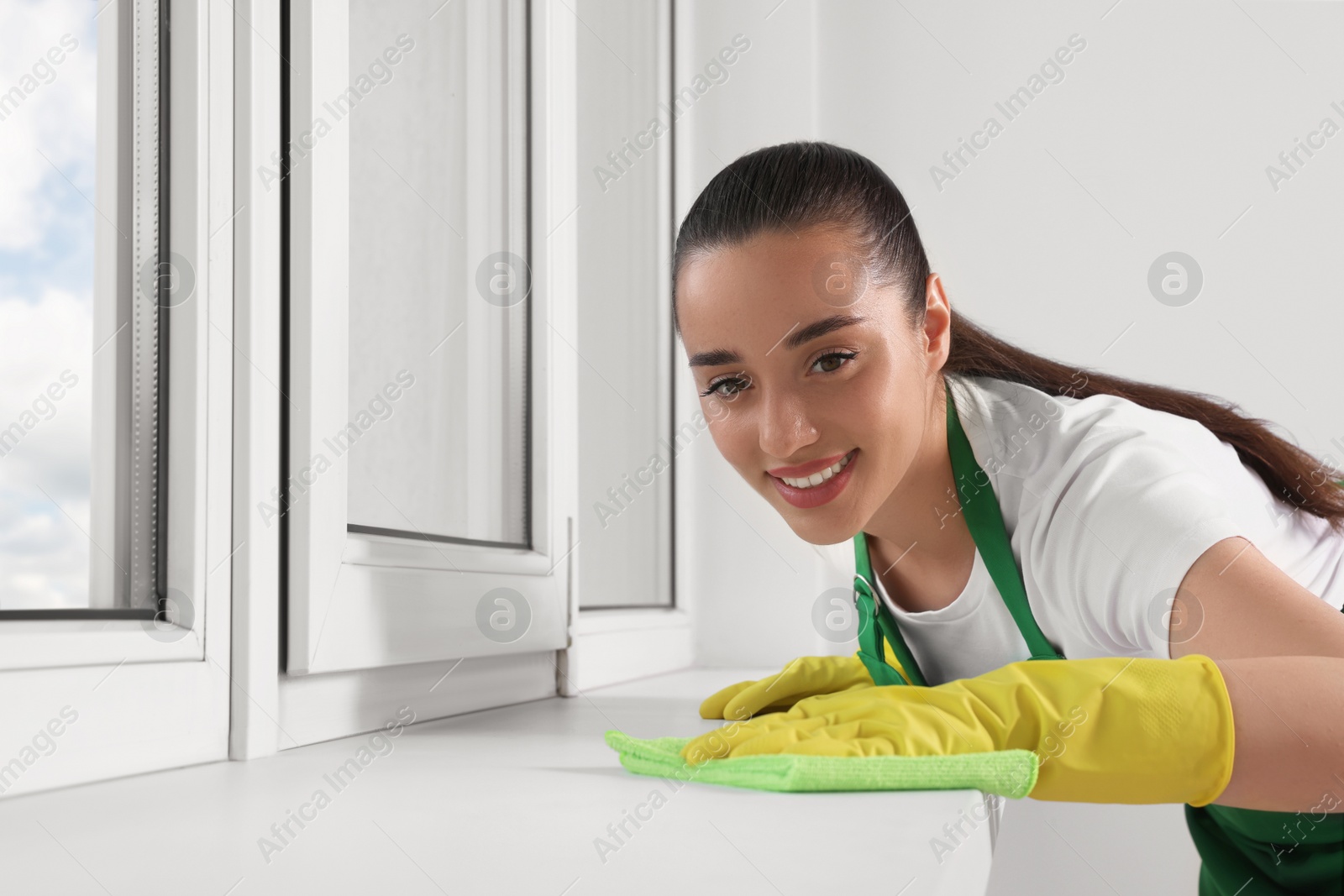 Photo of Happy young woman cleaning window sill with rag indoors