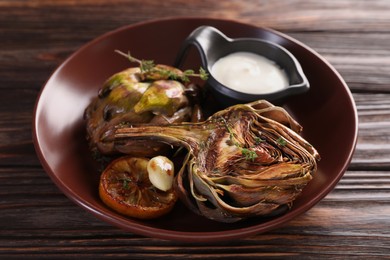 Photo of Tasty grilled artichoke and sauce in bowl on wooden table, closeup