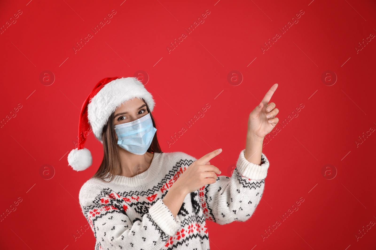 Photo of Pretty woman in Santa hat and medical mask pointing on red background