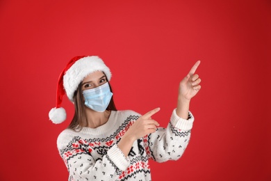 Photo of Pretty woman in Santa hat and medical mask pointing on red background