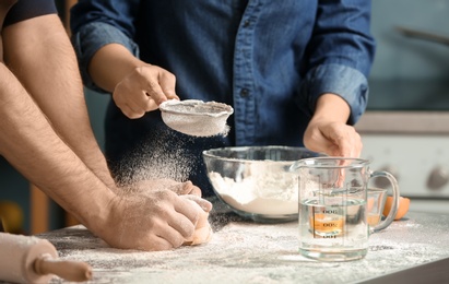 Photo of Woman sprinkling flour while man kneading dough on table in kitchen