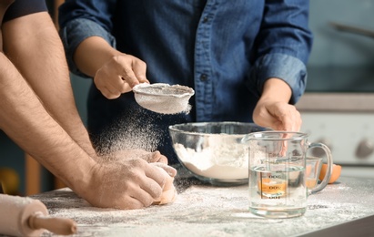 Woman sprinkling flour while man kneading dough on table in kitchen