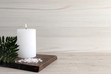 Burning candle, sea salt and green leaves on light beige wooden table, space for text