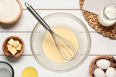 Photo of Glass bowl of crepe batter with whisk and ingredients on white wooden table, flat lay