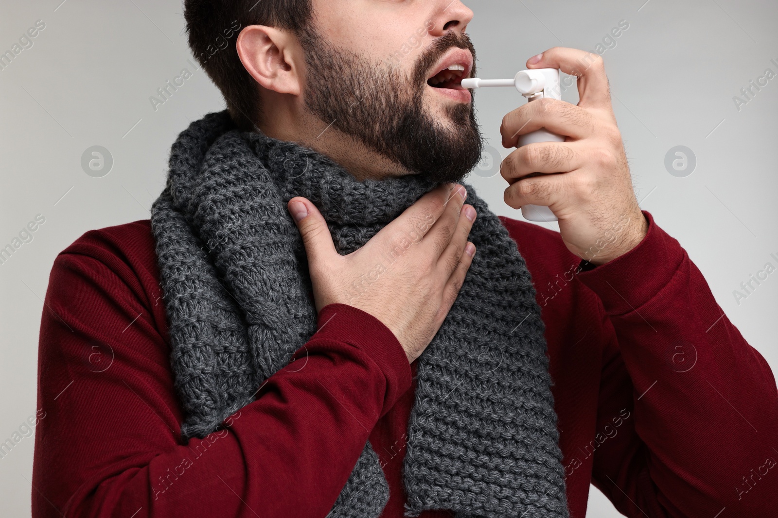 Photo of Young man with scarf using throat spray on grey background, closeup