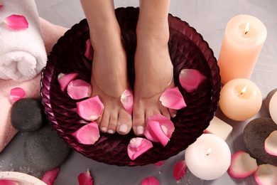 Photo of Woman soaking her feet in bowl with water and rose petals on grey floor, top view. Spa treatment
